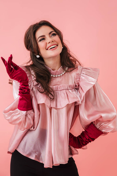 Portrait of a beautiful cheerful young brunette woman with bright makeup wearing fashionable clothes standing isolated over pink background, posing - 写真・画像