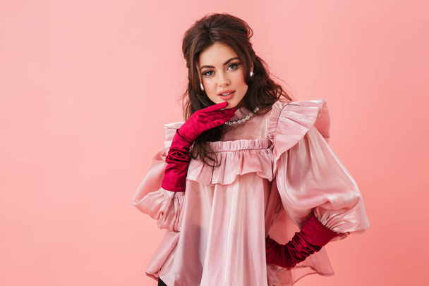 Portrait of a beautiful young brunette woman with bright makeup wearing fashionable clothes standing isolated over pink background, posing - 写真・画像
