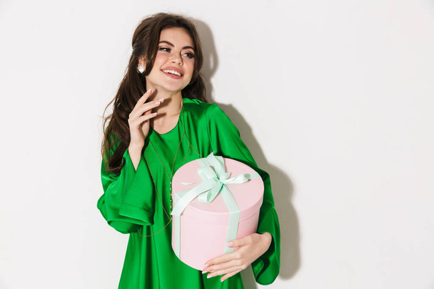 Portrait of a beautiful cheerful young brunette woman with bright makeup wearing fashionable clothes standing isolated over white background, holding present box - Fotoğraf, Görsel
