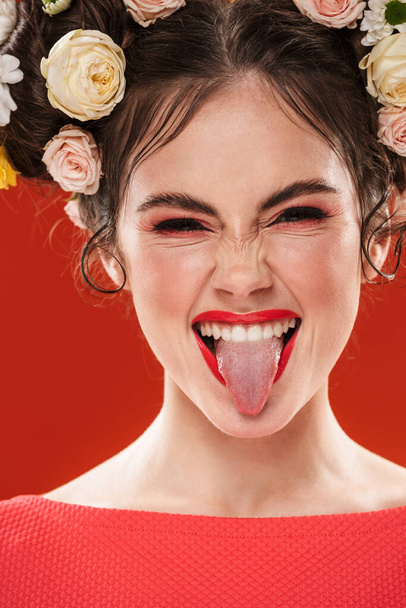Beautiful smiling young brunette woman with an amazing floral hairstyle wearing makeup posing isolated over red background, showing tongue - Foto, immagini