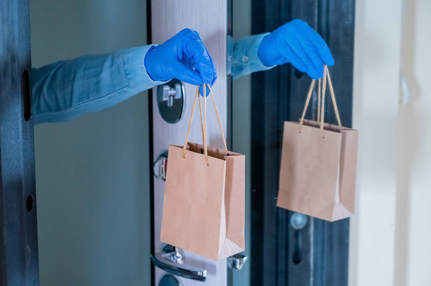 Close-up of a couriers hands in gloves with a paper bag in their hands at the front door. Quarantine delivery service. - Фото, изображение