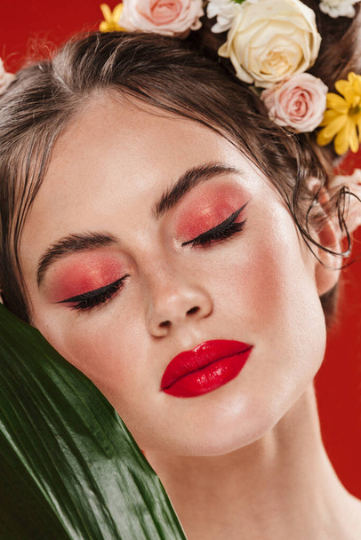 Krásná mladá brunetka žena s úžasnou květinový účes na sobě make-up pózovat s tropickým listem izolované přes červené pozadí - Fotografie, Obrázek