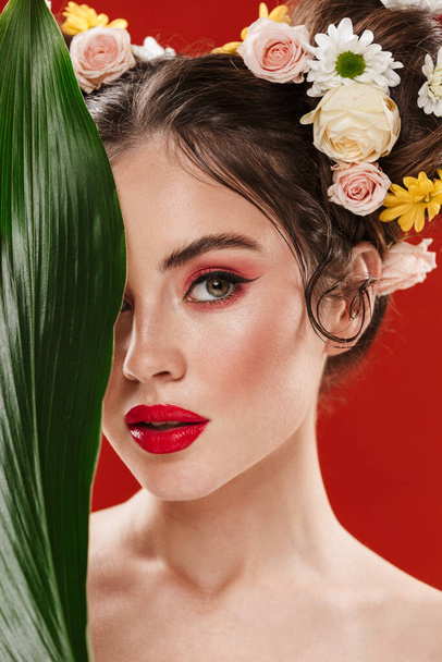 Beautiful young brunette woman with an amazing floral hairstyle wearing makeup posing with tropical leaf isolated over red background - 写真・画像