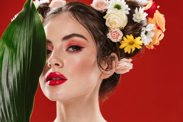 Beautiful young brunette woman with an amazing floral hairstyle wearing makeup posing with tropical leaf isolated over red background - Fotoğraf, Görsel