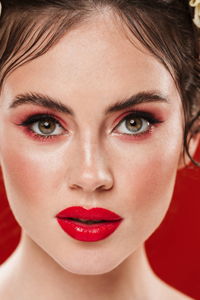 Close up of a beautiful young brunette woman wearing bright makeup isolated over red background, looking at camera - Zdjęcie, obraz