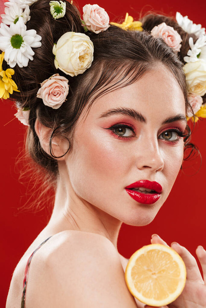 Beautiful young brunette woman with an amazing floral hairstyle wearing makeup posing with lemon isolated over red background - 写真・画像