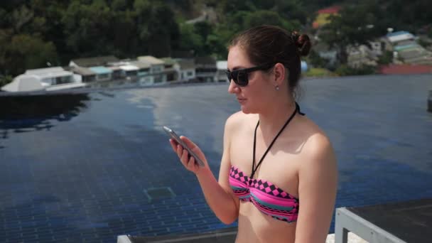 young woman types on black smartphone smiling near pool - Filmati, video