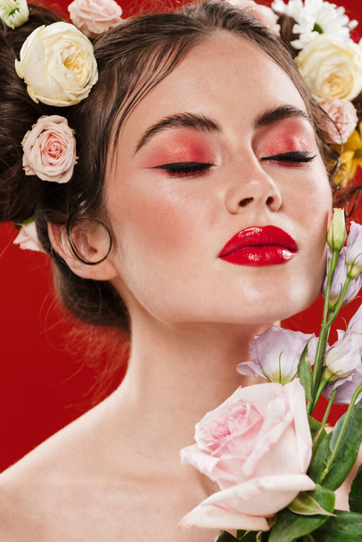 Close up portrait of a beautiful young brunette woman with a floral hairstyle and bright makeup posing isolated over red background, eyes closed, holding flowers - Photo, image