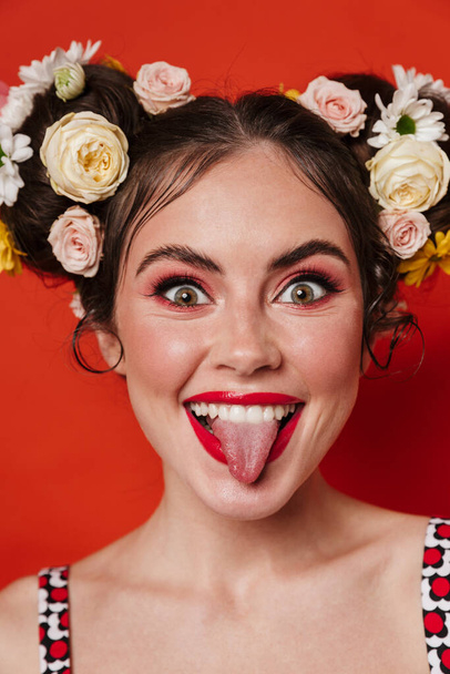 Close up portrait of a beautiful smiling young brunette woman with a floral hairstyle and bright makeup posing isolated over red background, grimacing - 写真・画像