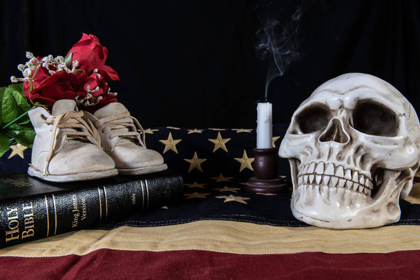 натюрморт, изображающий рождение и смерть нации в детской обуви на библейском и человеческом черепе и дымящейся свече на американском флаге
 - Фото, изображение