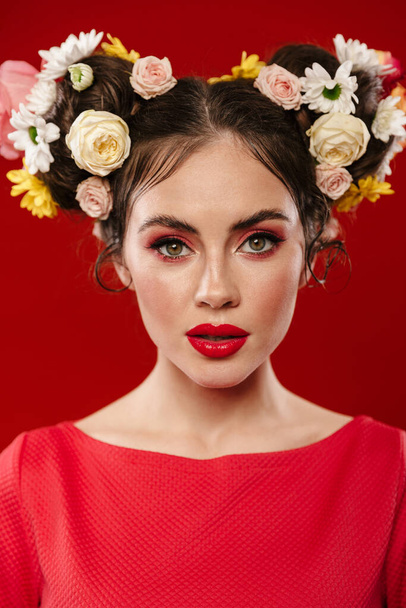 Beautiful young brunette woman with an amazing floral hairstyle wearing makeup posing isolated over red background, looking at camera - Фото, изображение