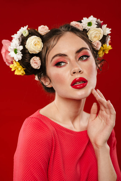 Beautiful young brunette woman with an amazing floral hairstyle wearing makeup posing isolated over red background - Valokuva, kuva