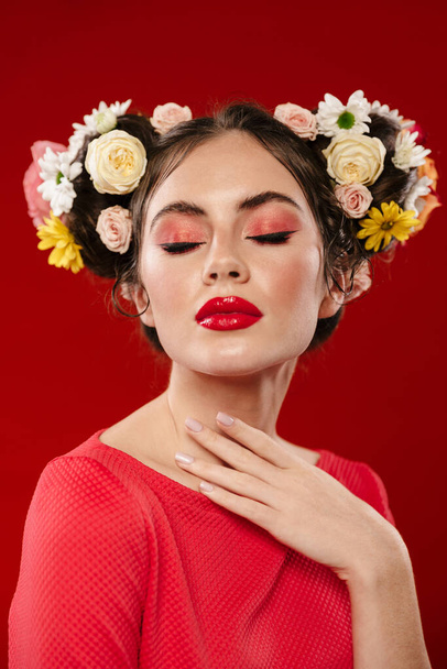Beautiful young brunette woman with an amazing floral hairstyle wearing makeup posing isolated over red background, eyes closed - Fotografie, Obrázek