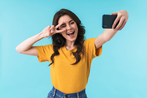 Image of happy positive young pretty woman posing isolated over blue wall background take selfie by mobile phone showing peace gesture. - Valokuva, kuva