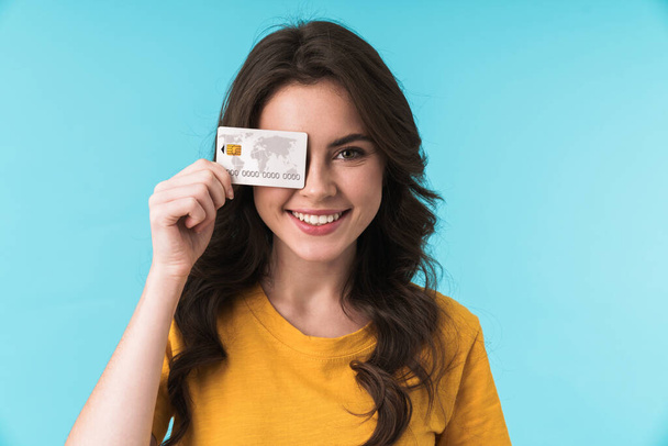 Image of emotional positive smiling young pretty woman posing isolated over blue wall background holding credit card covering eye. - Foto, Imagen
