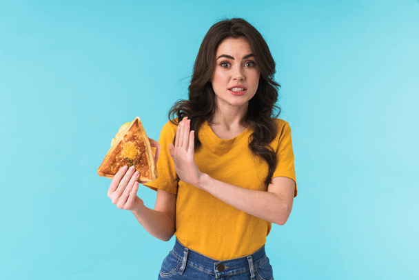 Image of displeased young beautiful woman posing isolated over blue wall background holding sandwich showing stop gesture. - Fotografie, Obrázek