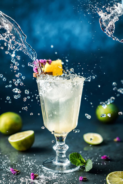 Φρέσκο κοκτέιλ λεμονάδας με λάιμ, μέντα και πάγο σε ποτήρι κρασιού σε σκούρο μπλε φόντο. Στιγμιότυπο από ποτό σε παγωμένη κίνηση, ιπτάμενες σταγόνες σε υγρό παφλασμό. Θερινό κρύο ποτό και κοκτέιλ - Φωτογραφία, εικόνα