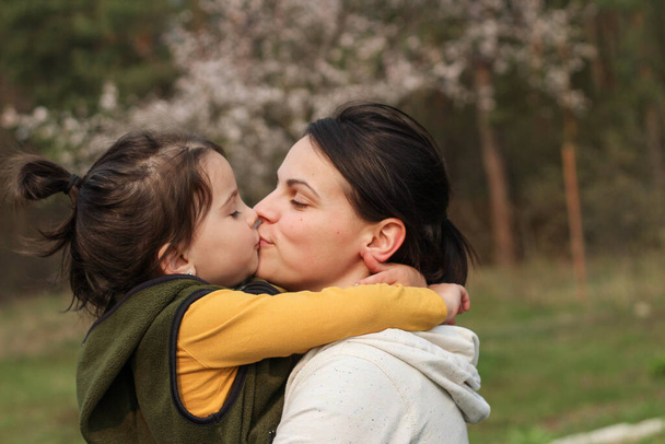 Gyönyörű anya és lánya ölelkeznek és csókolóznak alkalmi ruhákban. Meleg baba ölelés a háttérben egy elmosódott virágzó fa - Fotó, kép