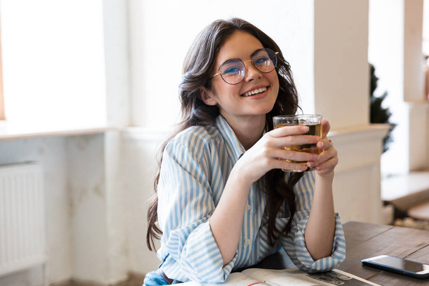 Close up of a smiling young brunette woman wearing shirt drinking juice while sitting in the cafe indoors - Φωτογραφία, εικόνα