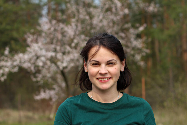 Een lachend mooi, schattig wit meisje met donker haar op de schouders staat in een groen T-shirt tegen de achtergrond van een wazige bloeiende lenteboom met bokeh. - Foto, afbeelding