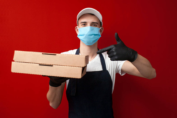 保護マスクと手袋を着た若いピザ配達人は箱とポイントを持っていますコロナウイルスの流行中の配達 - 写真・画像