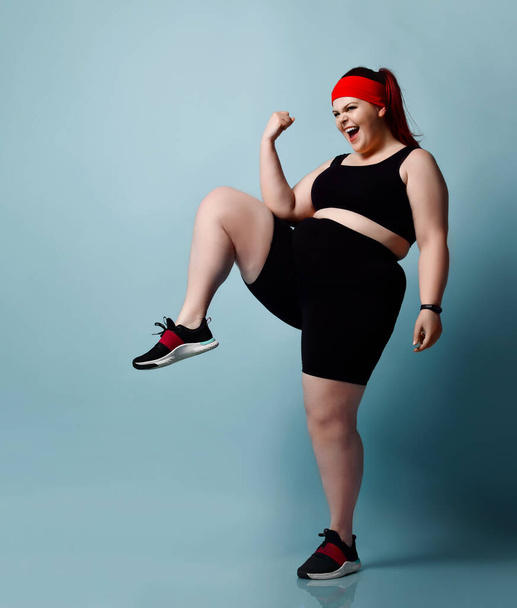 smiling plus-size girl practices exercises stepping in place raising up knees high on blue background. Fitness Full length portrait, copy space - Zdjęcie, obraz