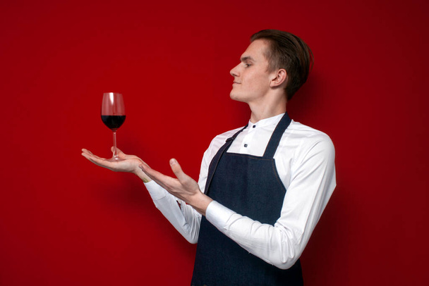 νεαρός επαγγελματίας σερβιτόρος με στολή κρατά ένα ποτήρι κόκκινο κρασί και δείχνει το χέρι του σε αυτό σε ένα κόκκινο φόντο, ένας σομελιέ τύπος συνιστά κρασί - Φωτογραφία, εικόνα