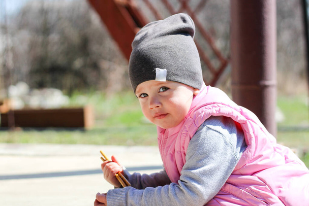 маленька біла дитина в сірому капелюсі і рожевий жилет накладає лікті на бетонну веранду на відкритому повітрі
 - Фото, зображення