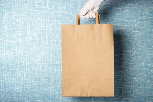 A woman's hand in a white glove holds cardboard bag, against a blue background - Photo, Image
