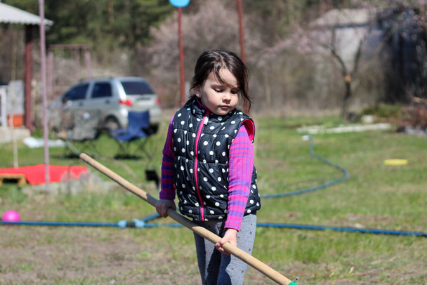a little white dark-haired white girl with a ponytail outdoors holds a long wooden stick in her hands. Preschooler helps parents in the spring garden - Photo, Image