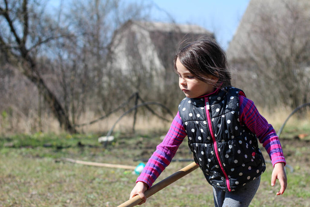 a little white dark-haired white girl with a ponytail outdoors holds a long wooden stick in her hands. Preschooler helps parents in the spring garden - Photo, image