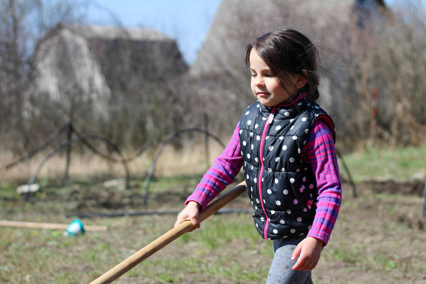 маленькая белая брюнетка с хвостиком на улице держит длинную деревянную палку в руках. Дошкольник помогает родителям в весеннем саду
 - Фото, изображение