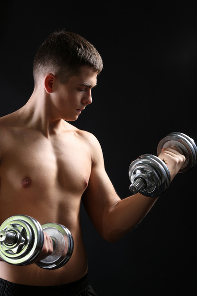 Handsome young muscular sportsman execute exercise with dumbbell on dark background - 写真・画像