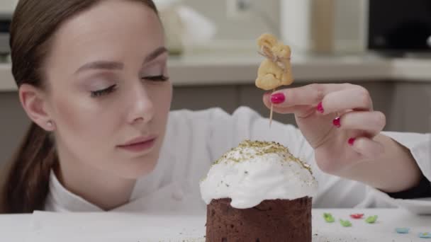ζαχαροπλάστης γυναίκα σε λευκό χιτώνα σε μια επαγγελματική κουζίνα, διακοσμεί κέικ Πάσχα, βάζει στην κορυφή μπισκότα σε σχήμα κουνελιού, χαμογελά - Πλάνα, βίντεο