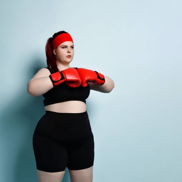 Close up portrait of concentrated young woman with overweight in red boxing gloves on blue background. Physical training, weight loss, martial arts - Photo, Image