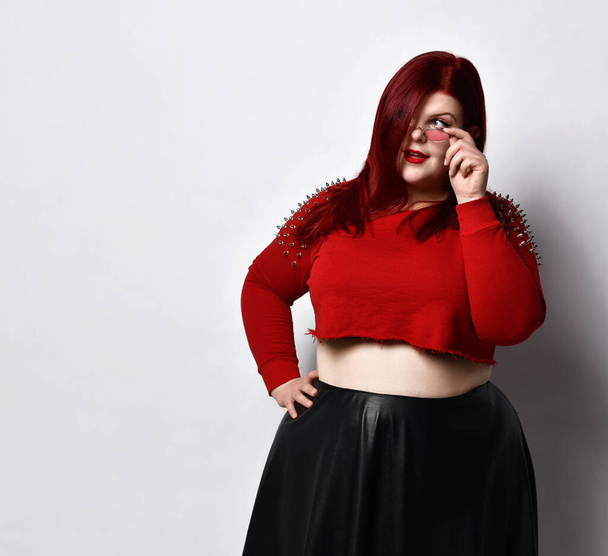 Overweight redhead woman in red spiked top, black skirt. She lowered her sunglasses and looking at you, posing isolated on white - Fotó, kép