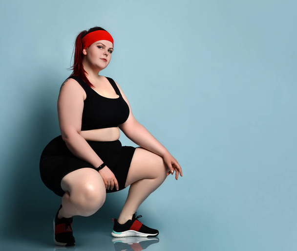 Modelo de jengibre con sobrepeso en diadema roja, top negro, shorts, zapatillas. Squatting posando de lado contra fondo azul del estudio
 - Foto, Imagen