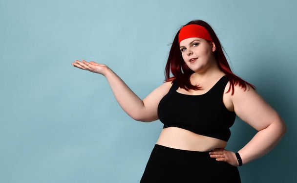 Overweight redhead female in red headband, black top and leggings. She holding something on her palm, posing on blue background - Fotó, kép