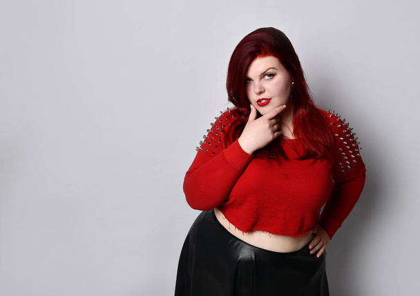 Obese redhead lady in red spiked top, black leather skirt, earrings. Touching face, put hand on waist, posing isolated on white - Photo, image