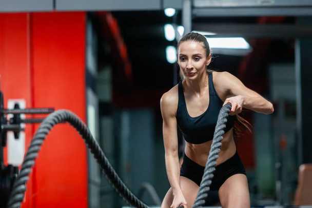 An attractive young muscular girl using training ropes for battle ropes exercise in a gym - Foto, immagini