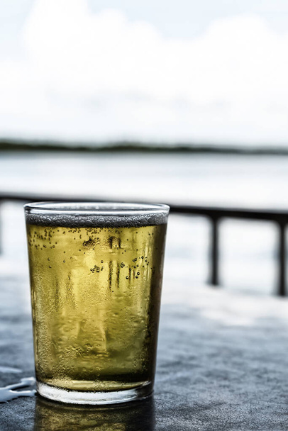 Glas helles Bier Pilsner steht auf dem Tisch eines Sommercafés auf dem verschwommenen Hintergrund des Flusses - Foto, Bild