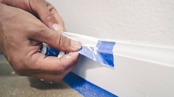 Removing masking tape from molding. A painter pulls of blue painter's tape from the wall to reveal a clean edge baseboard. - Foto, Imagen