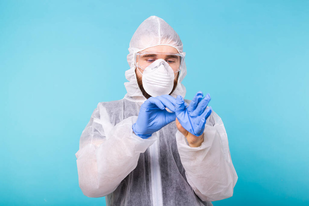 Mann in weißem Dekontaminationsanzug zieht medizinische Handschuhe an, isoliert auf blauem Hintergrund. Coronavirus, Covid-19 und Pandemiekonzept. - Foto, Bild