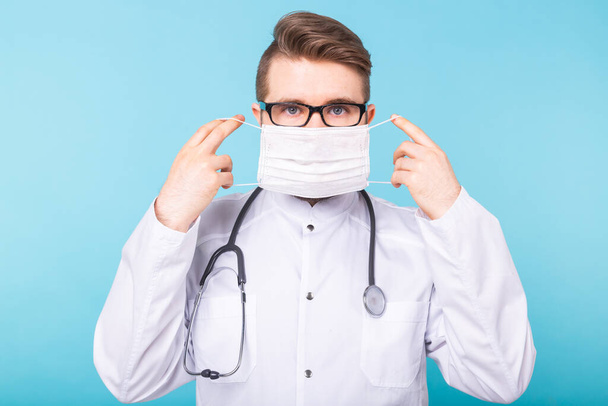 Muž doktor nasadí lékařskou masku na modré pozadí. Koronavirus, covid-19 a pandemická koncepce. - Fotografie, Obrázek