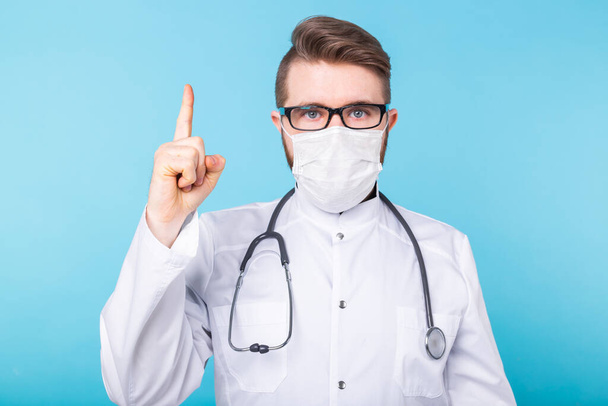 Portrét doktora v masce a bílé uniformě s prstem na modrém pozadí. Pozor, vypuknutí koronaviru, pandemie a kovid-19. - Fotografie, Obrázek