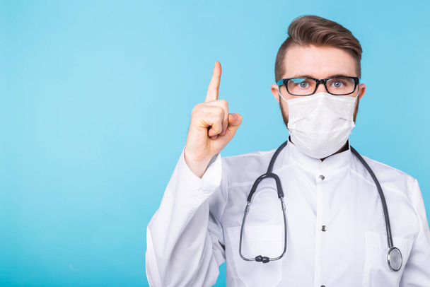 Retrato del médico con una máscara y uniforme blanco apuntando hacia arriba con un dedo sobre fondo azul con espacio para copiar. Atención, brote de coronavirus, pandemia y covid-19
. - Foto, imagen