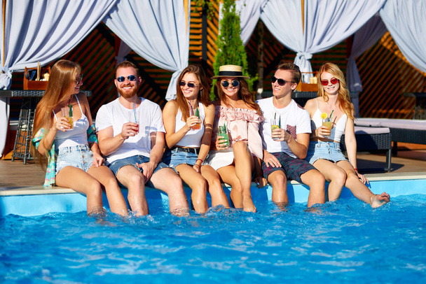 Group of friends having fun at poolside party clinking glasses with fresh cocktails sitting by swimming pool on sunny summer day. People toast drinking beverages at luxury villa on tropical vacation. - Foto, Imagen