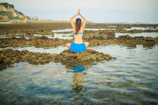 Young woman, meditating, practicing yoga and pranayama at the beach. Sunset yoga practice. Hands raising in namaste mudra. View from back. Water reflection. Melasti beach, Bali, Indonesia - Photo, Image