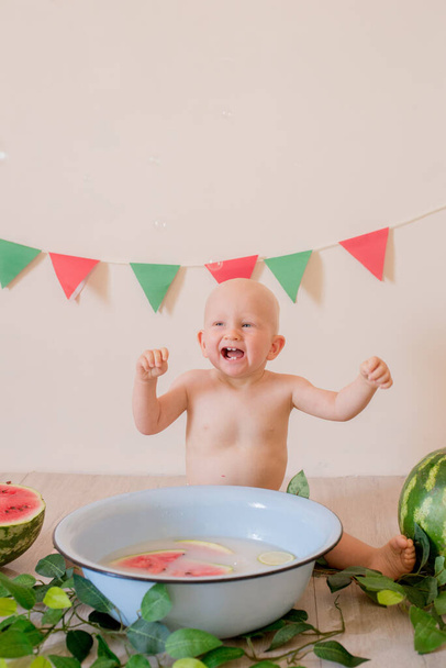 Little cute toddler with blond hair sits and splashes in a basin of water and watermelon on a  bright background. Children and fruits. Healthly food - Zdjęcie, obraz