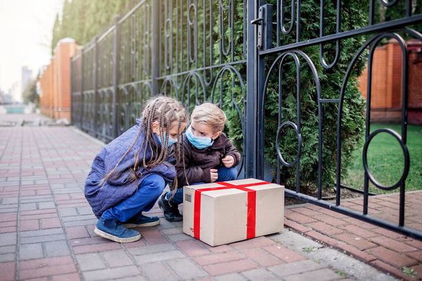 A kid wearing medical mask and gloves looking at delivered a cardboard box near the door of the modern house, outdoor lifestyle, self-isolation in lockdown, contactless online delivery - Photo, Image